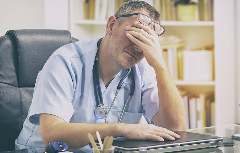 Foto eines Arztes am Schreibtisch in medizinischer Kleidung mit umgehängtem Stethoskop, der sich verzweifelt mit seiner linken Hand die Augen bedeckt