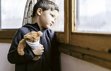 Foto eines kleinen Jungen, der traurig aus dem Fenster blickt. Im Arm hält er einen Teddybären, der eine Maske trägt.