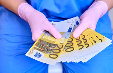 Foto von den Händen einer Pflegekraft im blauen Schutzkittel mit rosa Einmalhandschuhen. Sie halten ein Bündel 200 Euro-Scheine.