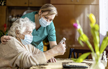 Foto von Pflegerin mit Maske, die einer sitzenden alten Dame - ebenfalls mit Maske - die Hand auf die Schulter legt und gemeinsam mit ihr das Etikett einer Medikamentenpackung studiert