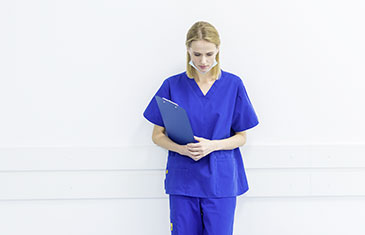 Foto einer jungen Pflegerin im dunkelblauen medizinischen Kittel, die mit einem Klemmbrett in der Hand sowie Maske unter dem Kinn kurz verschnauft