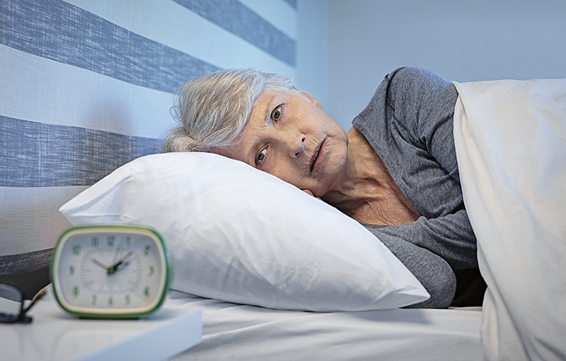 Foto einer älteren Frau, die wach im Bett liegt. Der Wecker zeigt kurz nach zwei Uhr an.