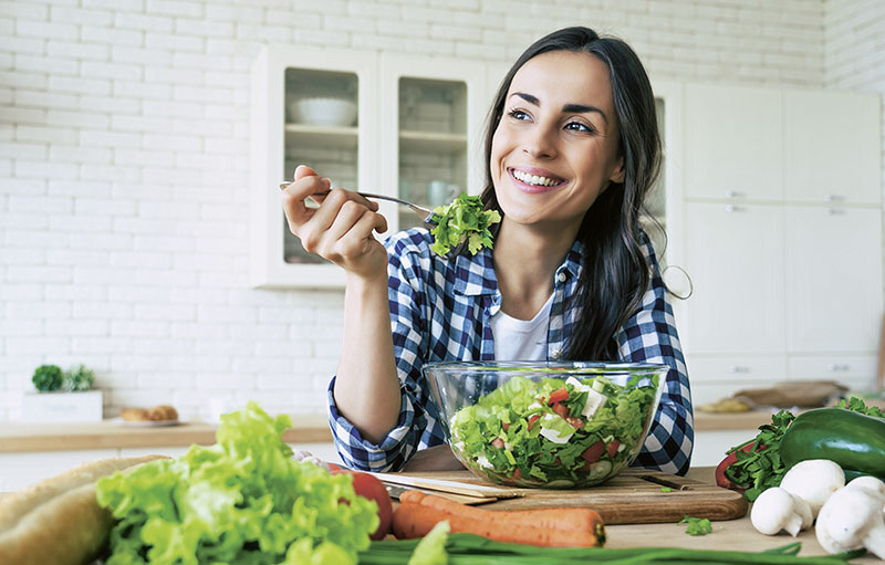 Foto einer jungen Frau, die gut gelaunt umgeben von Gemüse einen Salat isst