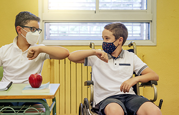Foto von zwei Jungen mit Maske in einem Unterrichtsraum. Einer von ihnen sitzt im Rollstuhl. Sie begrüßen sich mit dem Ellenbogen.