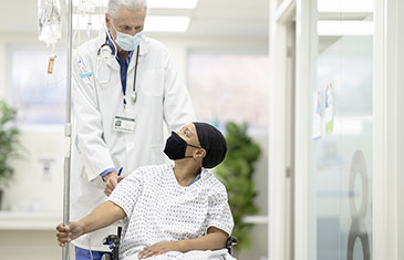 Foto einer Patientin mit Mütze, Kittel und Maske im Rollstuhl am Tropf. Sie wird von einem älteren Arzt mit Maske geschoben.