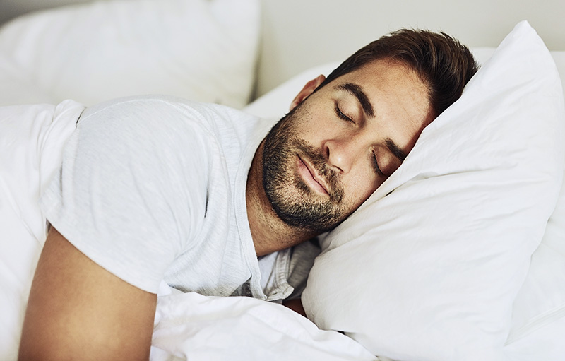 Foto eines jungen Mannes, der in weißem T-Shirt und weißer Bettwäsche seelig schläft