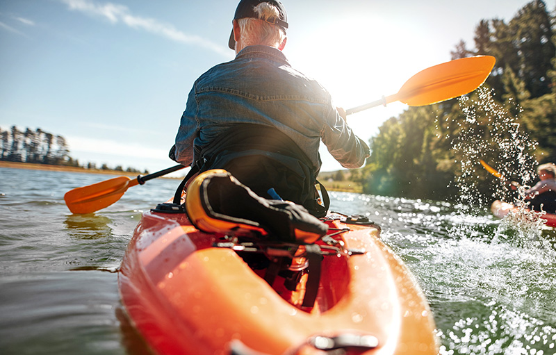 Foto eines älteren Mannes von hinten, der mit einem Kanu über einen See fährt. Rechts von ihm, am Rand des Bildes, ist eine Frau zu sehen, ebenfalls im Kanu.