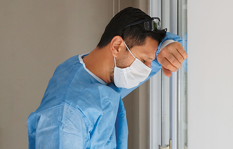 Foto einer männlichen Pflegekraft, die sich erschöpft mit Maske und blauem Kittel an die Wand lehnt