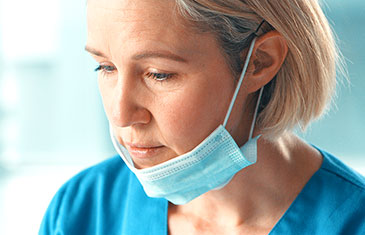 Foto einer weiblichen Pflegekraft mittleren Alters im blauen Arbeitskittel, die ihre Maske unter den Mund geschoben hat und müde aussieht