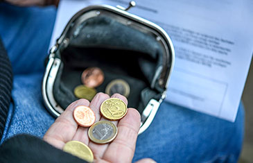 Foto einer offenen Geldbörse mit Kleingeld, das zusätzlich aus einer Hand in diese gegeben wird. Auf dem Schoß der Person liegt ein ausgedrucktes Dokument.