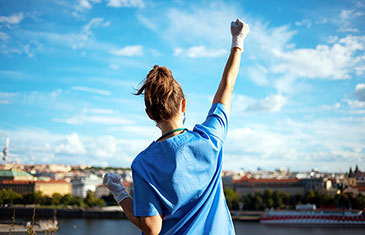 Foto einer Pflegerin im hellblauen Kittel, die über eine Stadtkulisse blickt und ihren rechten Arm in Siegerpose in die Höhe reckt