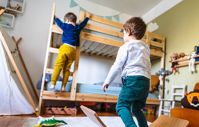 Foto von zwei Kleinkindern im Kinderzimmer. Das eine steigt die Treppe eines Hochbetts hoch. Das andere balanciert auf einem Balanceboard.