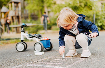 Foto eines kleinen Kindes, das mit bunter Kreide auf eine Straße malt. Im Hintergrund steht sein Dreirad.