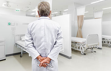 Foto eines Arztes von hinten im weißen Kittel mit Stethoskop, der in einem Mehrbettzimmer mit leeren Betten steht