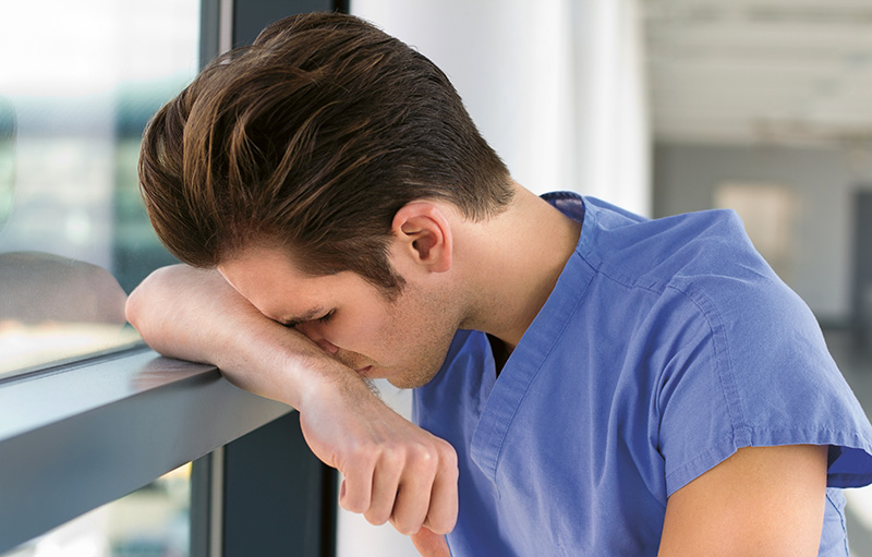 Foto eines jungen Pflegeschülers, der sich im blauen Kittel erschöpft an ein Fenster leht