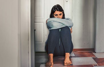 Foto einer jungen Frau, die mit verschränkten Armen traurig auf dem Boden vor einer Tür sitzt