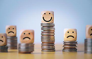Foto von mehreren Münzgeldstapel mit jeweils einem Smiley-Würfel obenauf, der mal lächelt, mal traurig dreinblickt