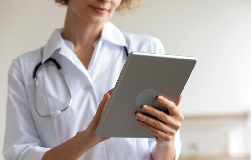 Symbolbild einer Ärztin in Arbeitskleidung mit Stethoskop, die ein Tablet hochkant in Händen hält, auf das sie blickt.