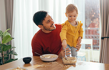 Symbolbild eines Vaters, der mit seinem Kleinkind zusammen Müsli isst. Das kleine Mädchen schaufelt Müsli in die Schüssel und kleckert dabei ein bisschen.