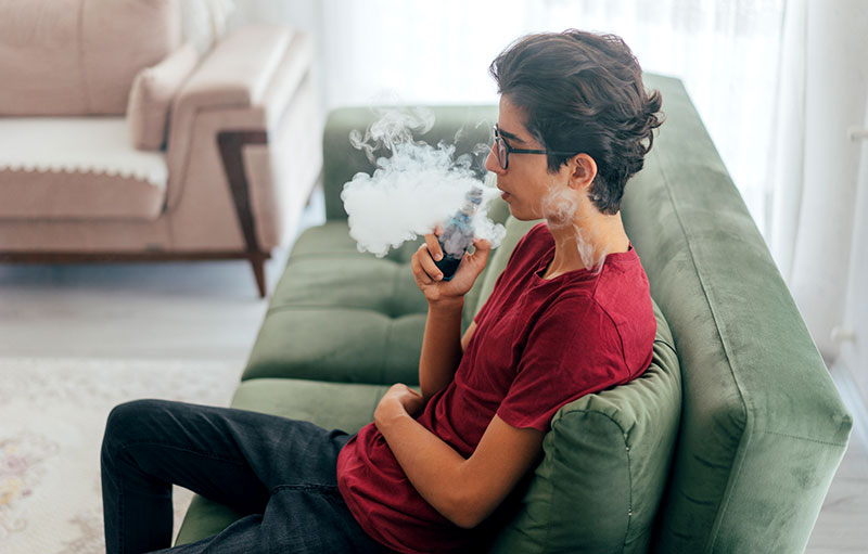 Symbolfoto eines mit einem Verdampfer rauchenden Jungen auf einem grünen Sofa