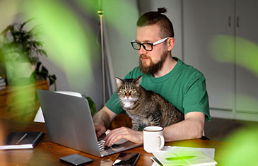 Foto von jungem Hipster mit Katze auf den Schoß, der im Homeoffice am Laptop sitzt