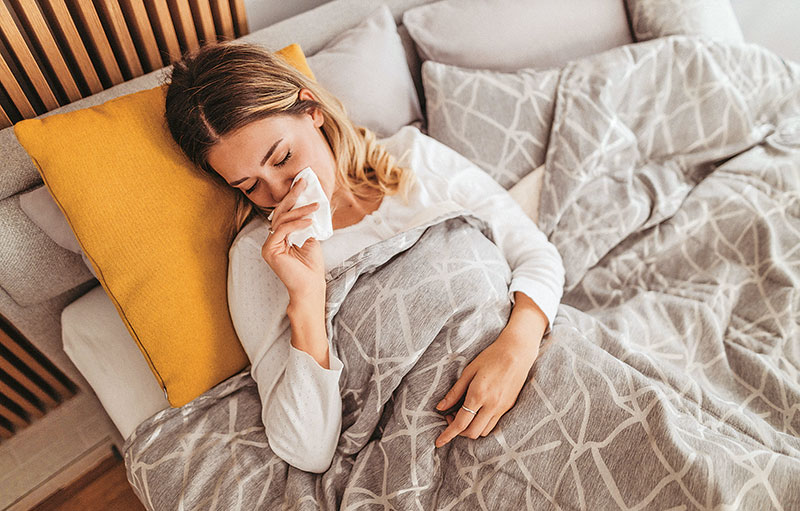 Symbolbild einer jungen Frau, die krank im Bett liegt und sich ein Taschentuch unter die Nase hält.
