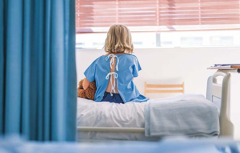 Symbolbild eines Kindes von hinten mit Teddy auf einem Krankenhausbett. Es trägt ein hinten offenes Leibchen.