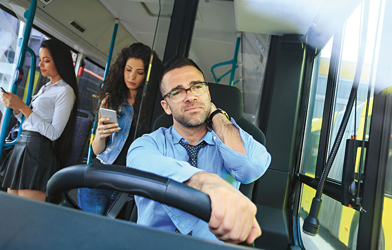 Foto eines Busfahrers, der sich den Nacken hält. Im Hintergrund sind Fahrgäste zu sehen.
