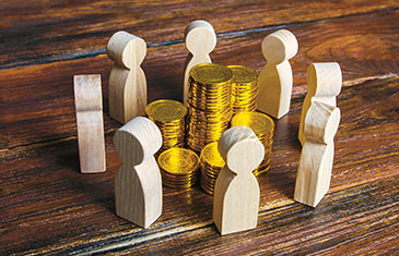 Symbolbild: Mehrere helle Holzfiguren in Menschenform stehen auf einem dunklen Holztisch im Kreis um mehrere Stapel Goldmünzen herum
