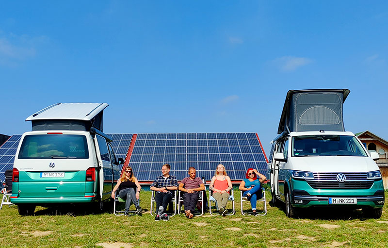 Foto von Julia, Christoph, Christoph, Jacqueline und Charis zwischen ihren beiden Vans auf Campingstühlen bei einer Rast. Hinter ihnen sind Solarpanele zu sehen.