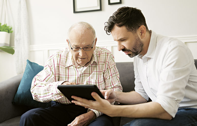 Foto eines Mannes mittleren Alters, die mit einem alten Mann zusammen auf dem Sofa sitzend in ein Tablet blickt