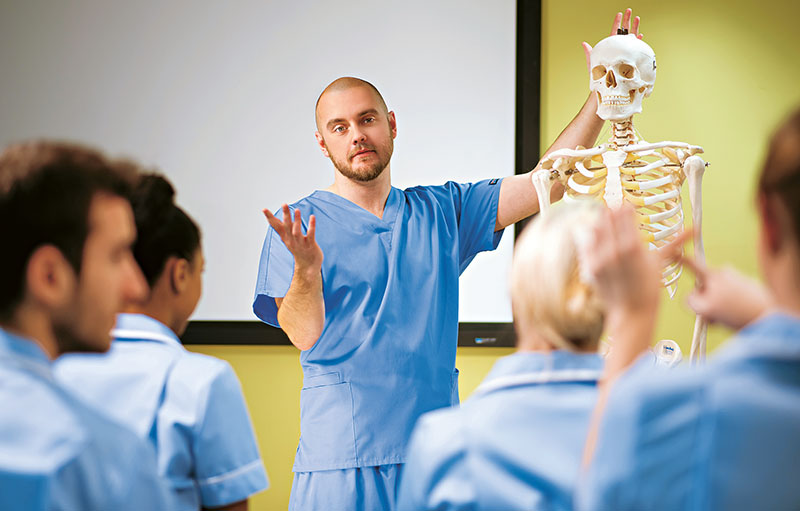 Foto von Auszubildenden mit blauen Kitteln in einem Seminarraum. Ein Lernender erklärt etwas an einem Skelett-Modell.