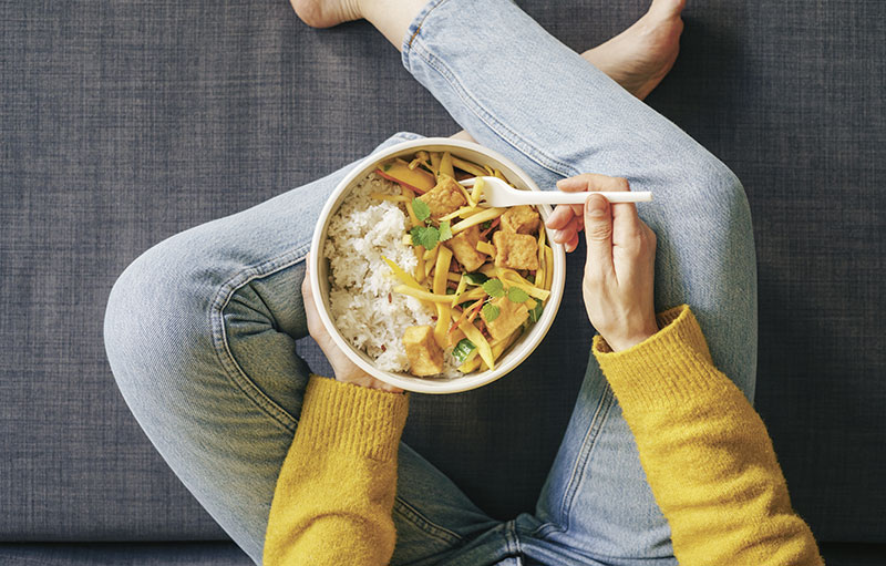 Foto einer Person, die im Schneidersitz auf dem Sofa sitzt und ein asiatisches, gelbes Gemüse-Curry isst