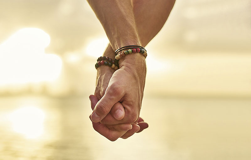 Foto von zwei Händen mit bunten Armbändern, die sich halten. Im Hintergrund ist verschwommen ein See im abnehmenden Sonnenlicht zu sehen.