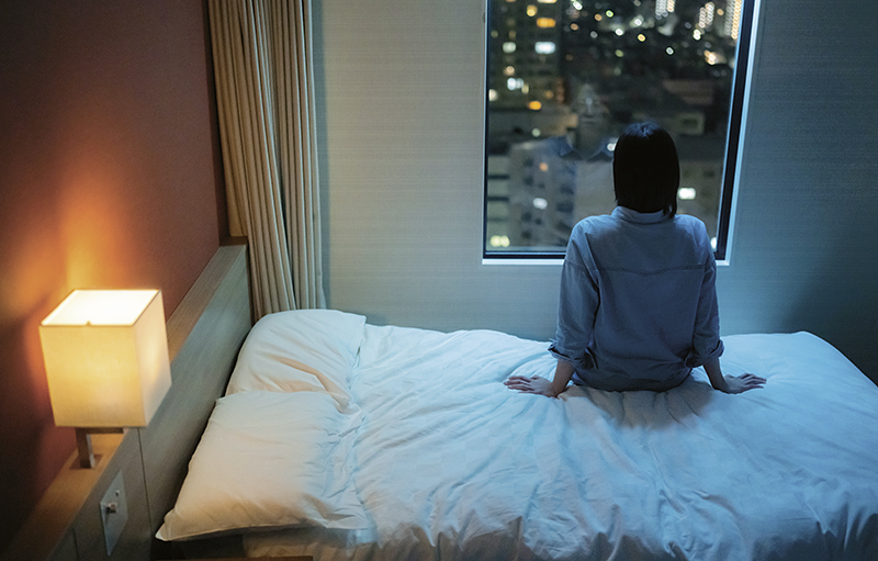 Foto einer jungen Frau, die bei Dunkelheit von einem Schlafzimmer aus auf eine Stadtkulisse blickt